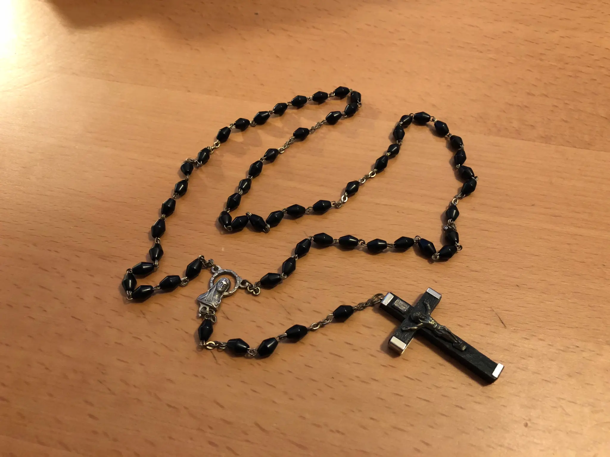 A black rosary made of faceted glass beads on a small silver chain, ending in a Mary medallion and a wood-and-metal crucifix, photographed on a wooden tabletop in warm available light.