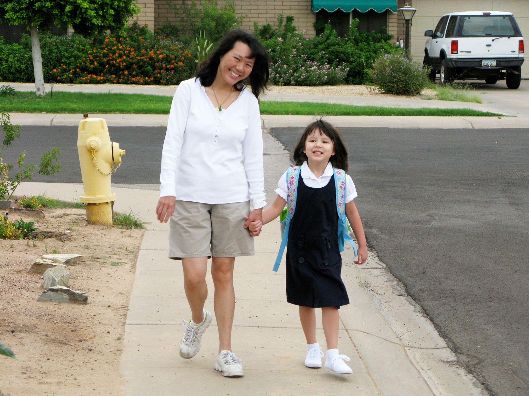 A young Morgan Kubasko, hair in pigtails, walks to her first day of kindergarten on a Tempe sidewalk holding her mother Margie's hand. A yellow fire hydrant and orange lantana flowers are in the background; a Ford Explorer is parked in a neighboring driveway.