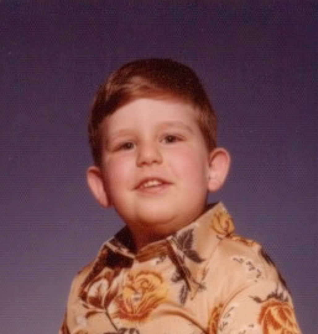 A studio school portrait of Rob Kubasko at approximately age four, c. 1976, wearing a floral shirt against a violet backdrop, smiling slightly with red hair neatly combed.