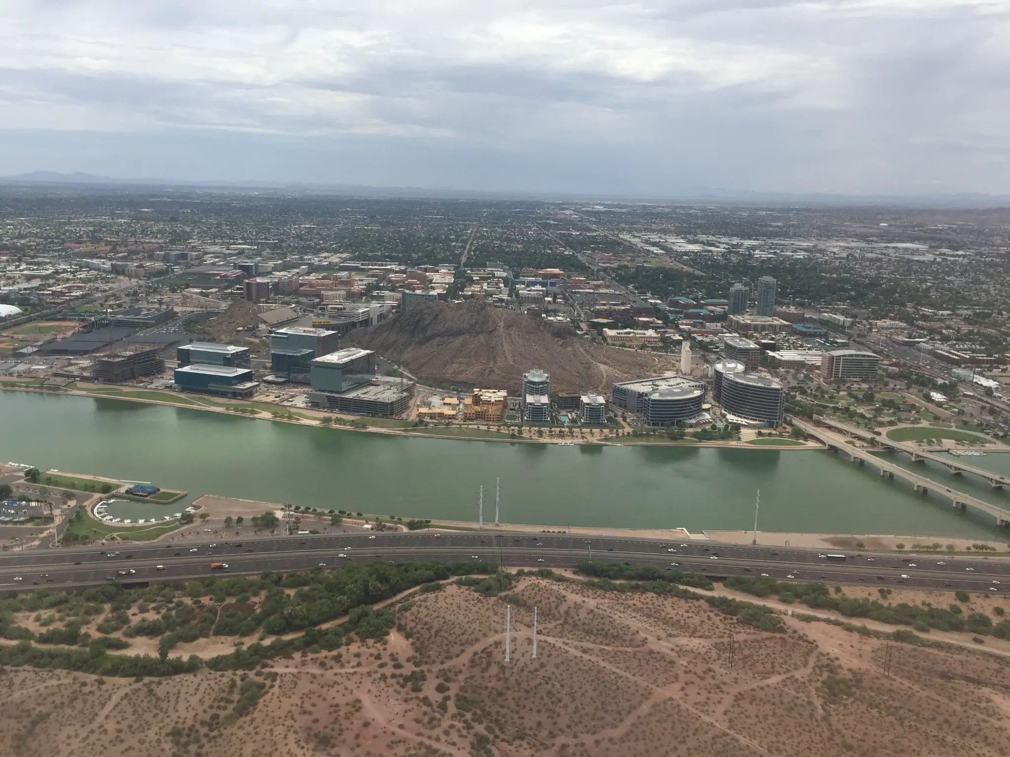 An aerial photograph of Tempe, Arizona, taken from a passenger window on descent. Town Lake curves across the frame, with Tempe Butte at center and Sun Devil Stadium and the ASU campus visible to the left.